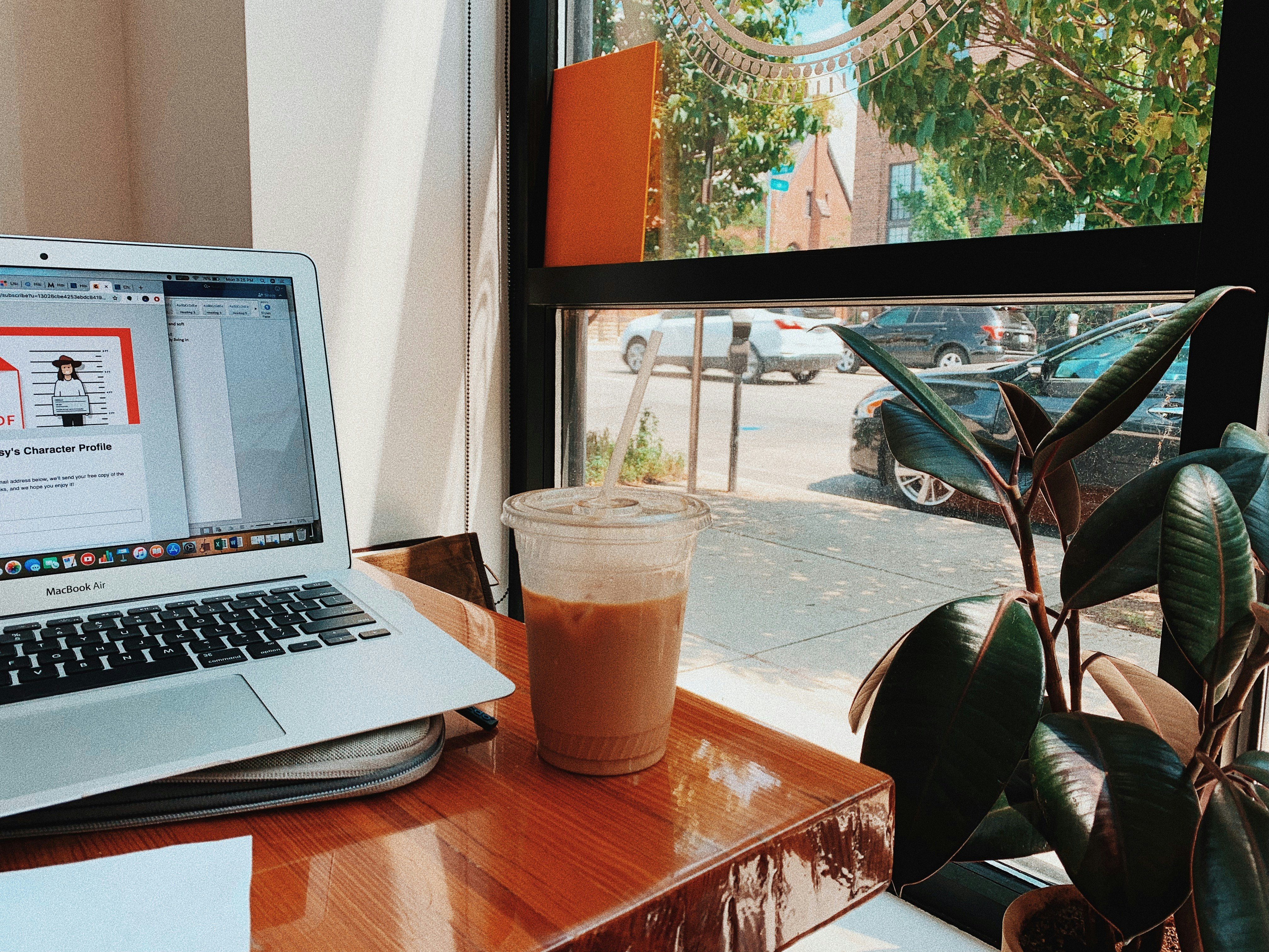 MacBook Air beside filled with plastic cup on table near rubber plant during daytime